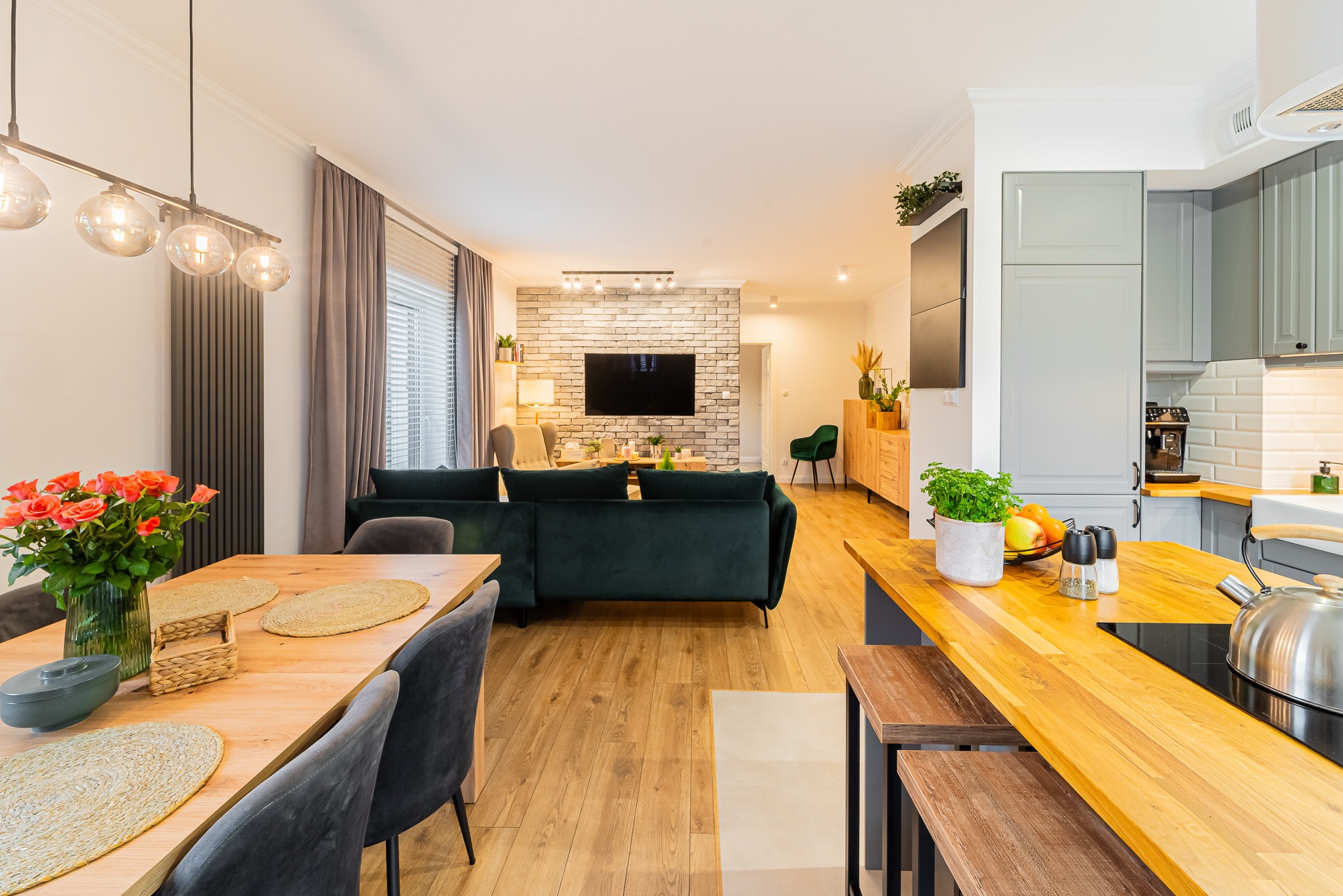 Przestronne wnętrze domu z otwartą kuchnią, jadalnią i salonem. Widoczny stół z kwiatami, zielona sofa i ceglana ściana z telewizorem. Nowoczesny design i jasne oświetlenie.