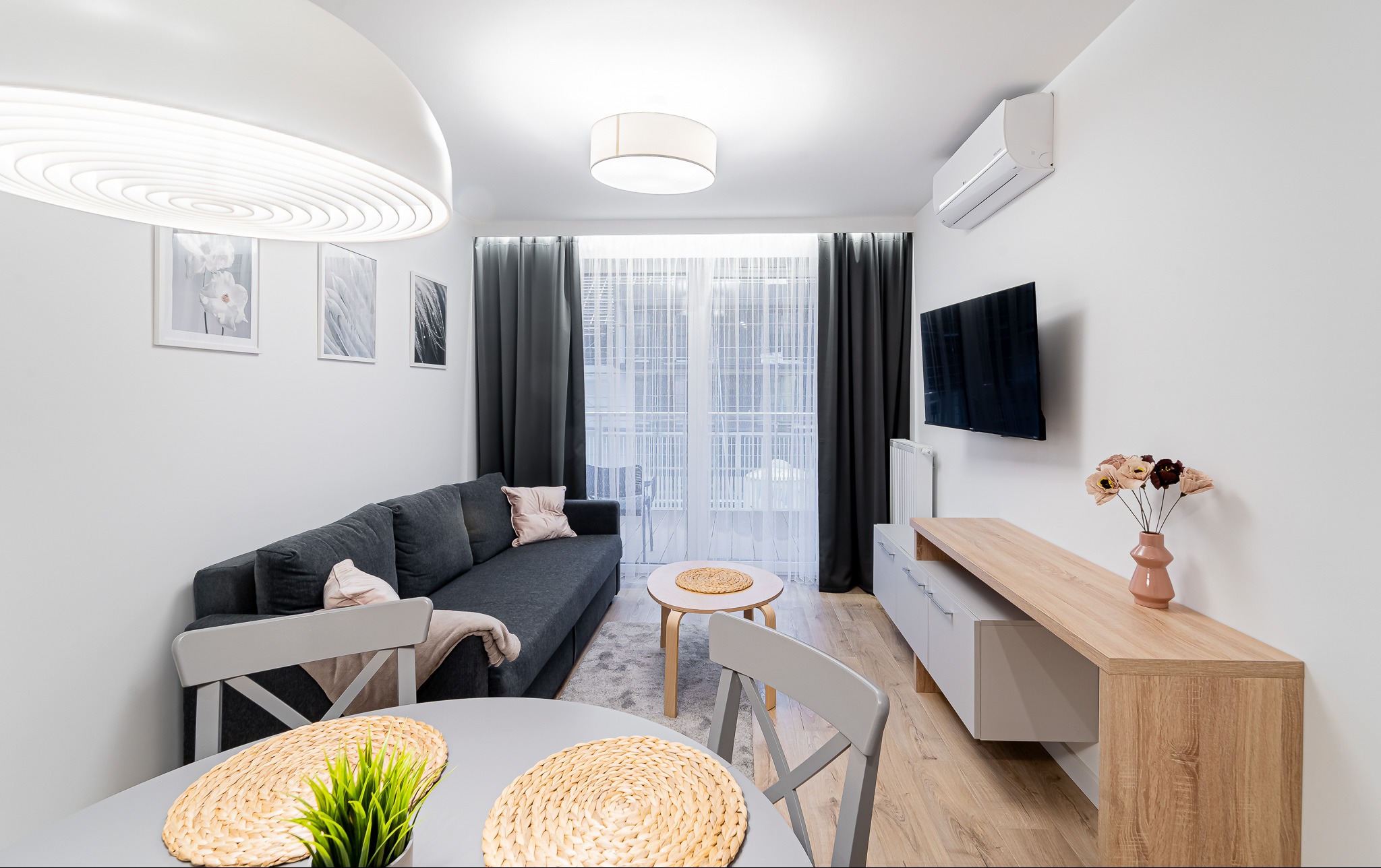 Jasne wnętrze salonu z szarą sofą, drewnianym stolikiem kawowym i telewizorem na ścianie. Widoczne okno z zasłonami i element jadalni z okrągłym stołem.