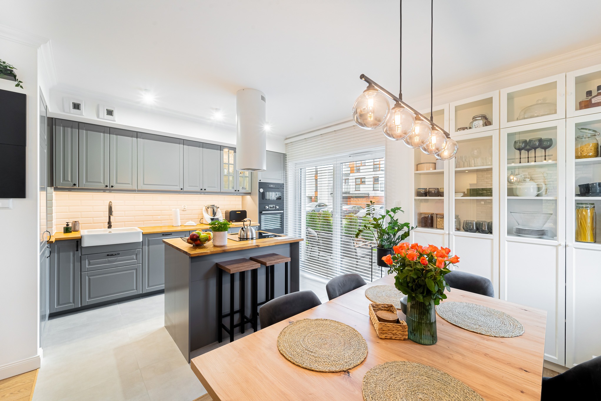 Jasna kuchnia w szarościach z drewnianym blatem i wyspą. Na stole bukiet pomarańczowych róż. Widoczne przeszklone szafki i nowoczesne oświetlenie.