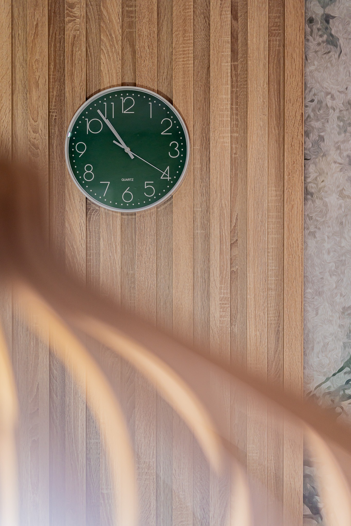 Zielony zegar ścienny na drewnianej boazerii, z delikatnym wzorem tapety w tle i elementem balustrady w dolnej części kadru. Minimalistyczny design wnętrza.
