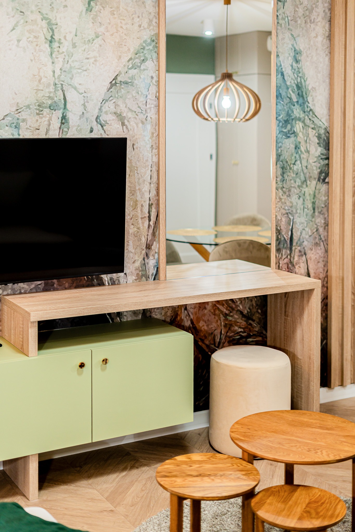 Nowoczesny salon z zieloną szafką RTV, drewnianym blatem i okrągłymi stolikami kawowymi. W tle lustro odbija lampę i stół. Tapeta z motywem roślinnym.