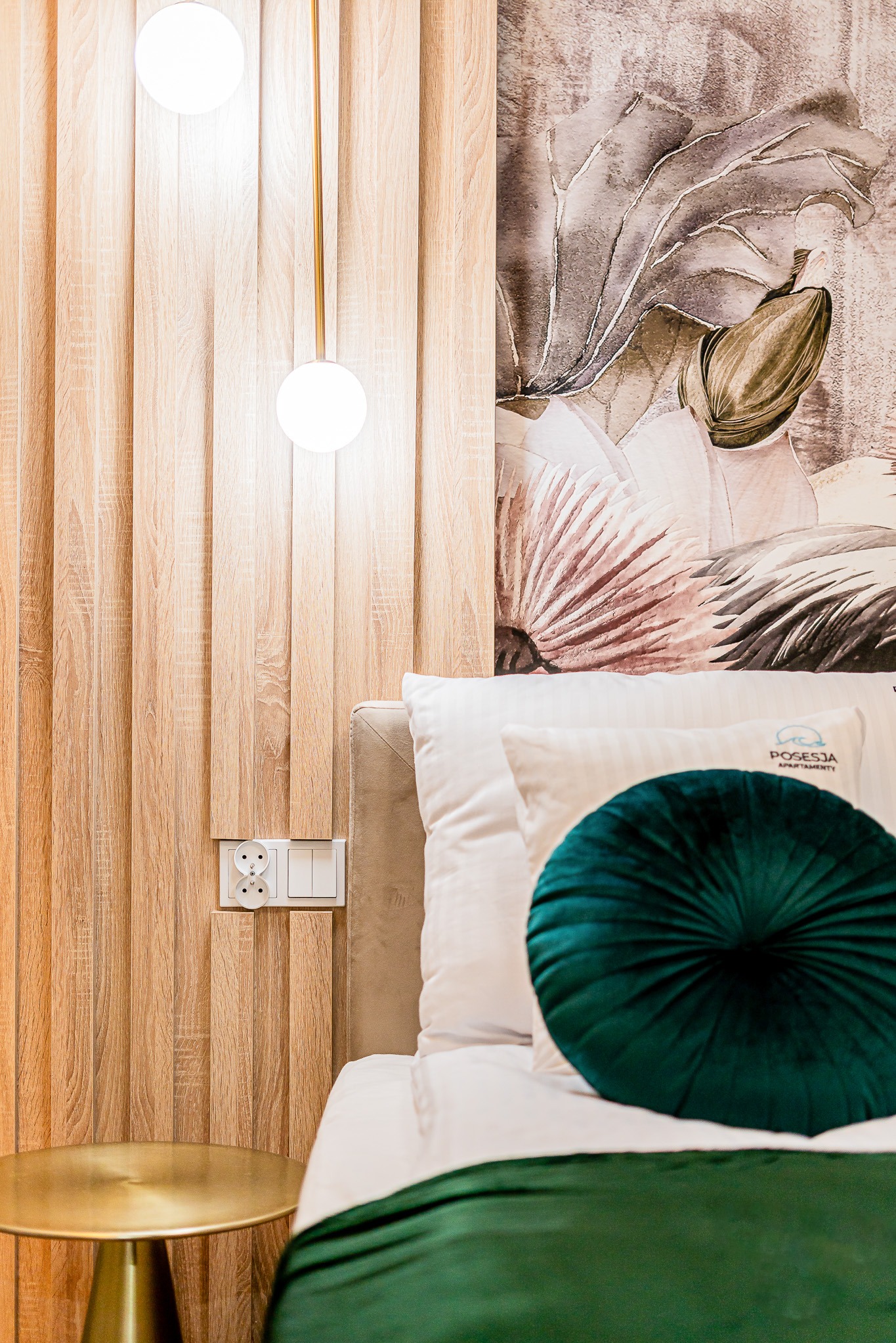 Fragment sypialni z drewnianą ścianą, lampą wiszącą, tapetą w kwiaty i łóżkiem z zieloną poduszką. Widoczne gniazdka elektryczne i stolik nocny.