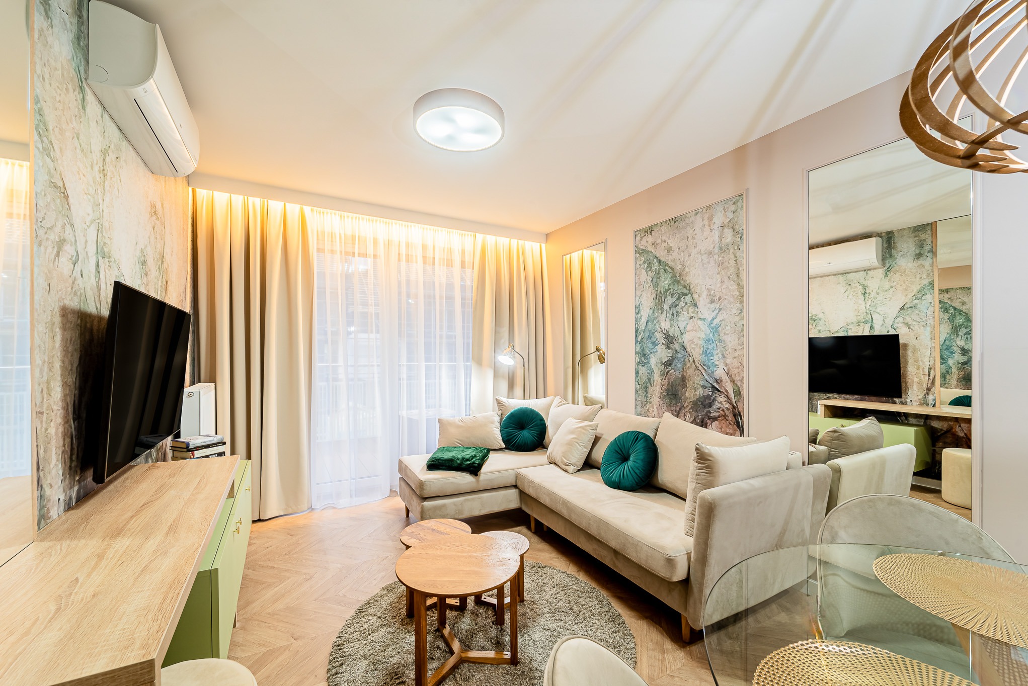 Jasne wnętrze salonu z narożną sofą, okrągłymi stolikami kawowymi i parkietem w jodełkę. Na ścianie abstrakcyjny obraz i lustro powiększające przestrzeń.