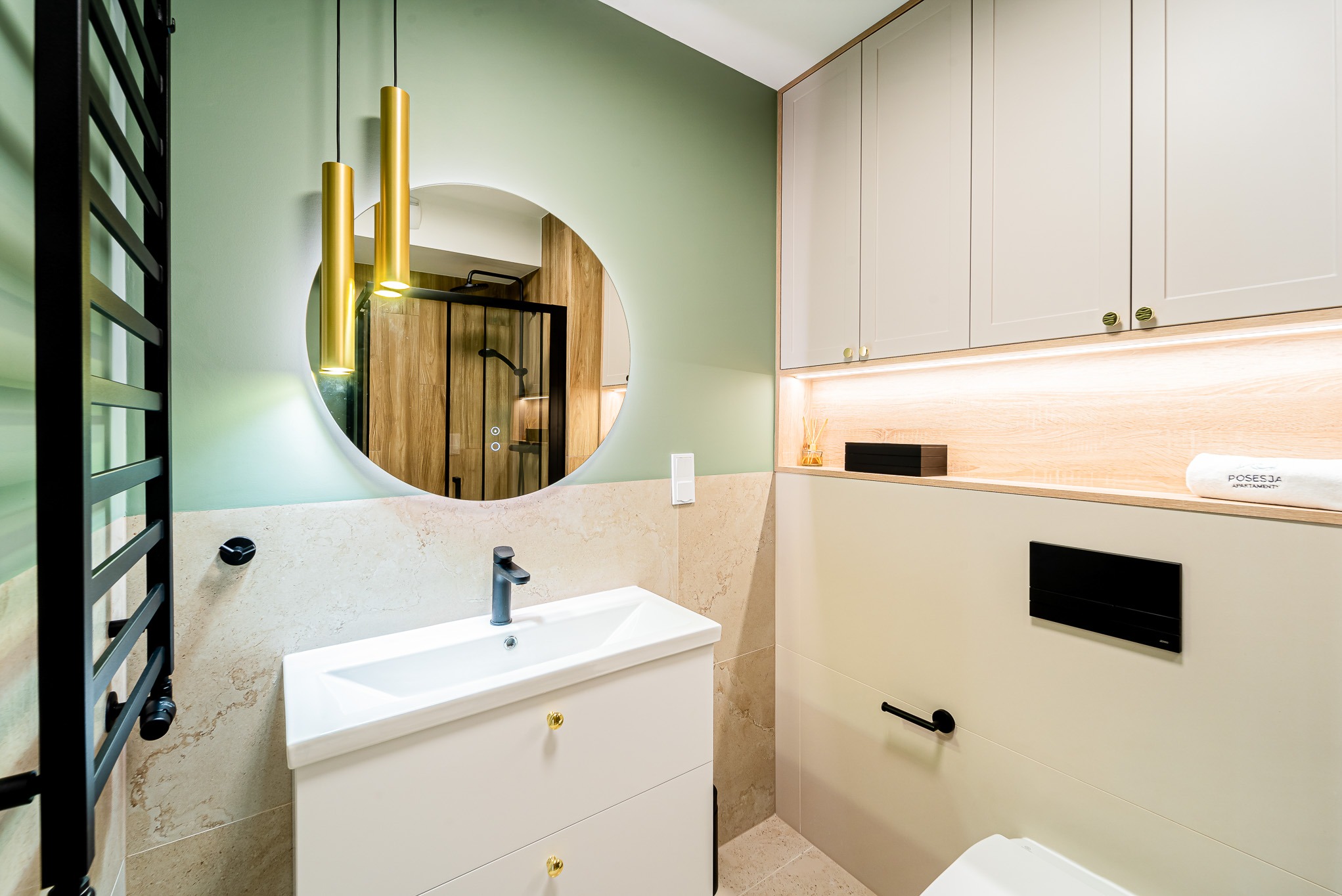 Nowoczesna łazienka z okrągłym lustrem podświetlanym LED, umywalką na szafce z złotymi uchwytami i czarnym grzejnikiem drabinkowym na tle pistacjowej ściany.