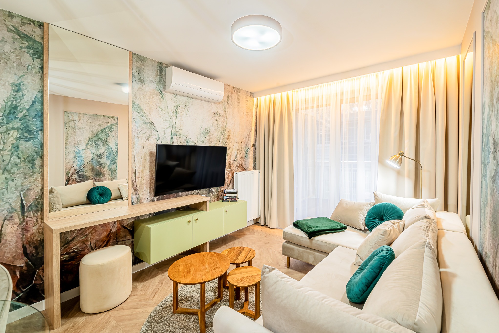 Jasne wnętrze salonu z telewizorem, kanapą i stolikami kawowymi. Ściana z wzorem przypominającym marmur, lustro i elementy dekoracyjne w odcieniach zieleni i beżu.