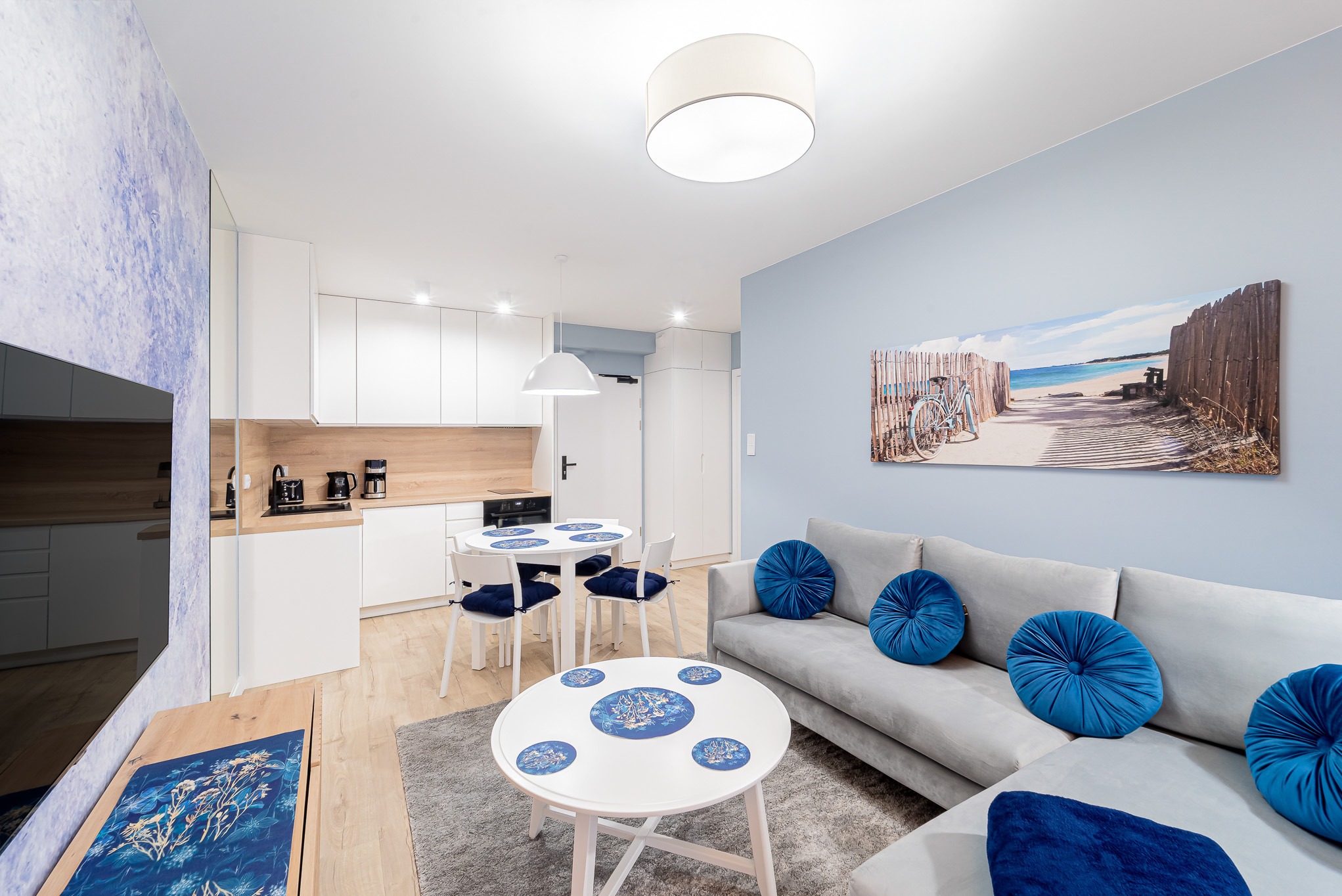 Jasne wnętrze mieszkania z aneksem kuchennym, okrągłymi stolikami i szarą sofą z niebieskimi poduszkami. Na ścianie obraz z motywem plaży i rowerem.