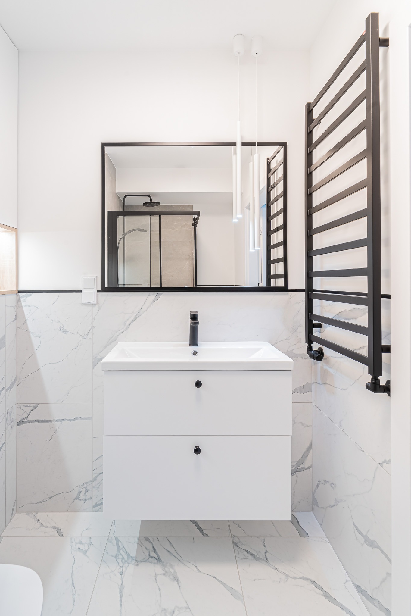 Nowoczesna łazienka z białymi marmurowymi płytkami, białą szafką pod umywalką i czarną armaturą. W lustrze odbija się kabina prysznicowa.