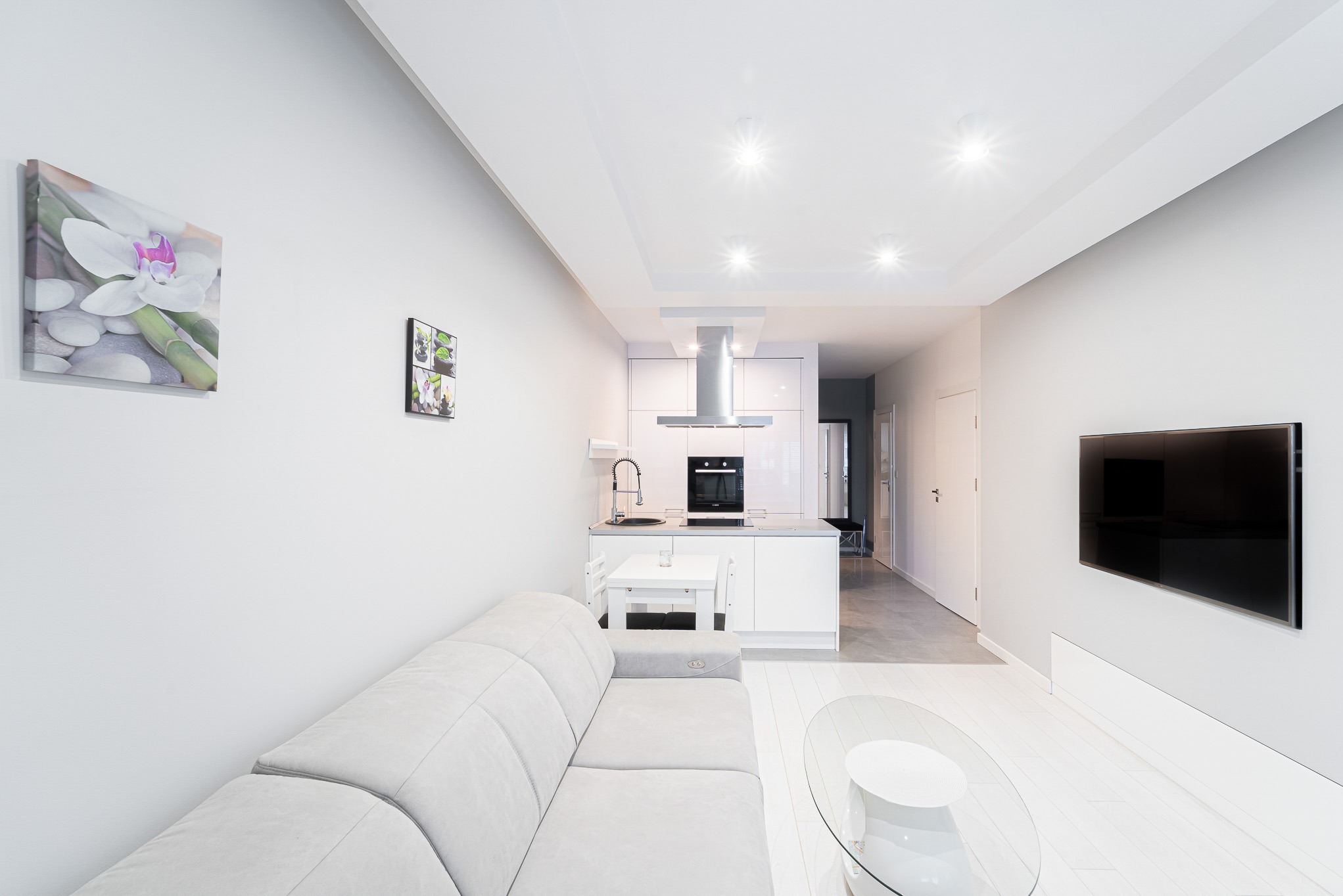 Jasne wnętrze apartamentu w Kołobrzegu: salon z sofą, aneks kuchenny z wyspą, minimalistyczny design, obrazy na ścianach, telewizor. Nowoczesne oświetlenie.