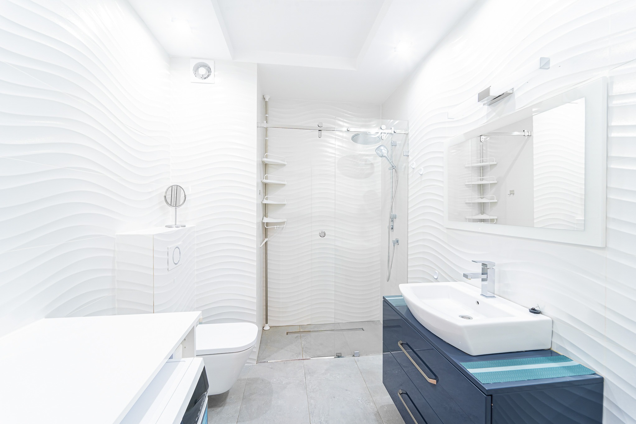 Nowoczesna, jasna łazienka z białą armaturą i ścianami z falistych płytek. Umywalka na szafce w kolorze granatowym. Kabina prysznicowa ze szklanymi drzwiami.