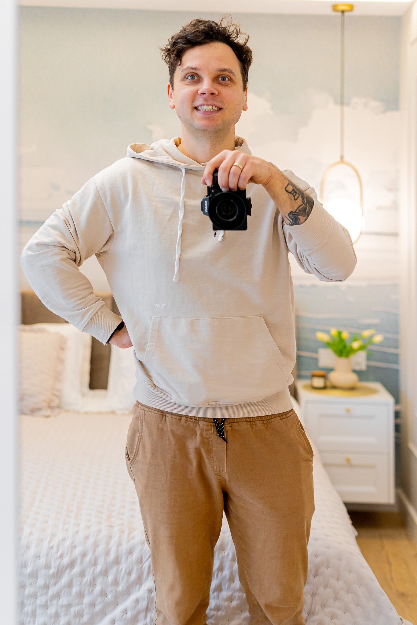 Uśmiechnięty mężczyzna w beżowej bluzie z kapturem, robiący sobie zdjęcie w lustrze, w tle widoczny pokój z łóżkiem i kwiatami.