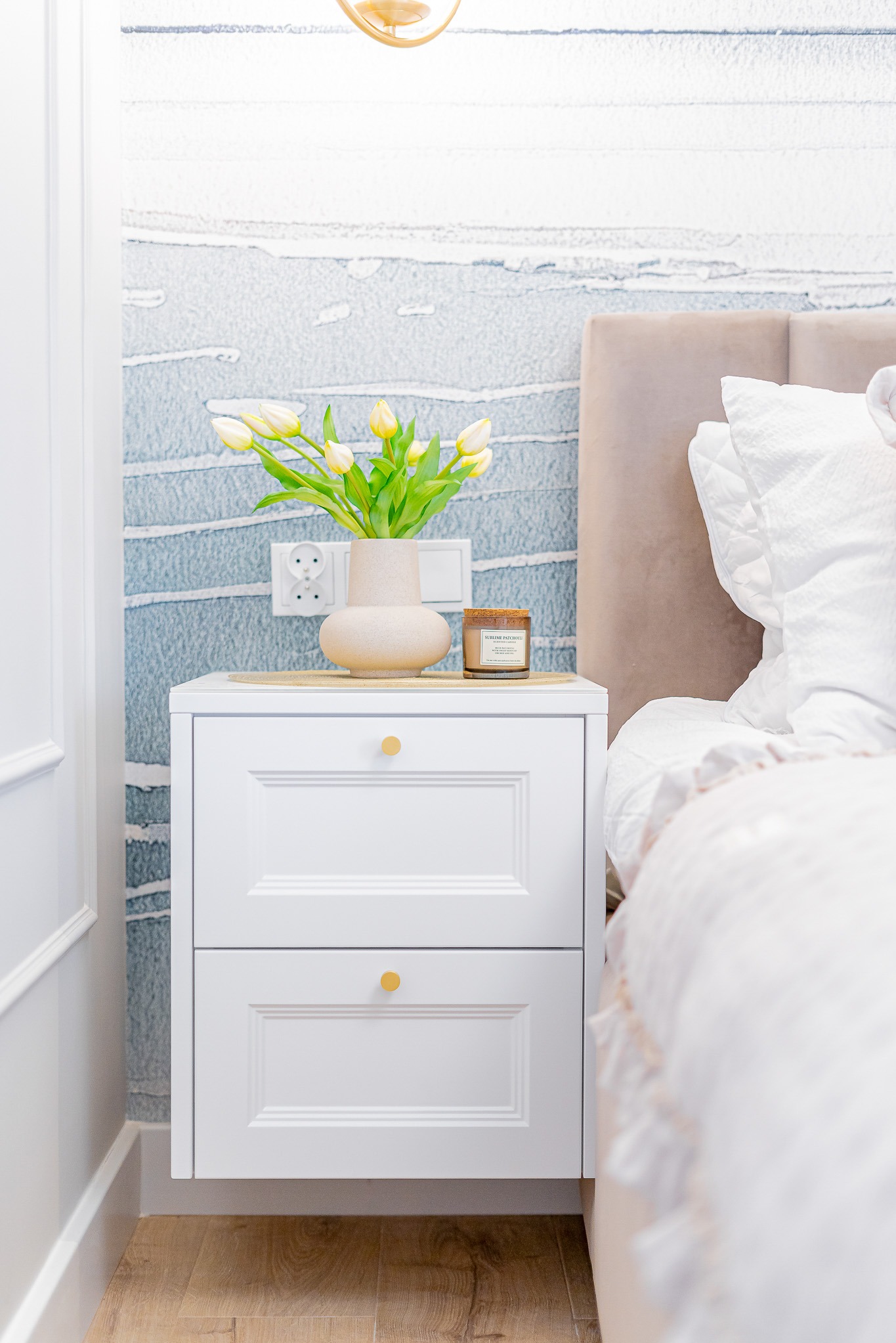 Biała szafka nocna z dwiema szufladami i złotymi uchwytami, na której stoi wazon z żółtymi tulipanami i świeca, na tle ściany z abstrakcyjnym wzorem.