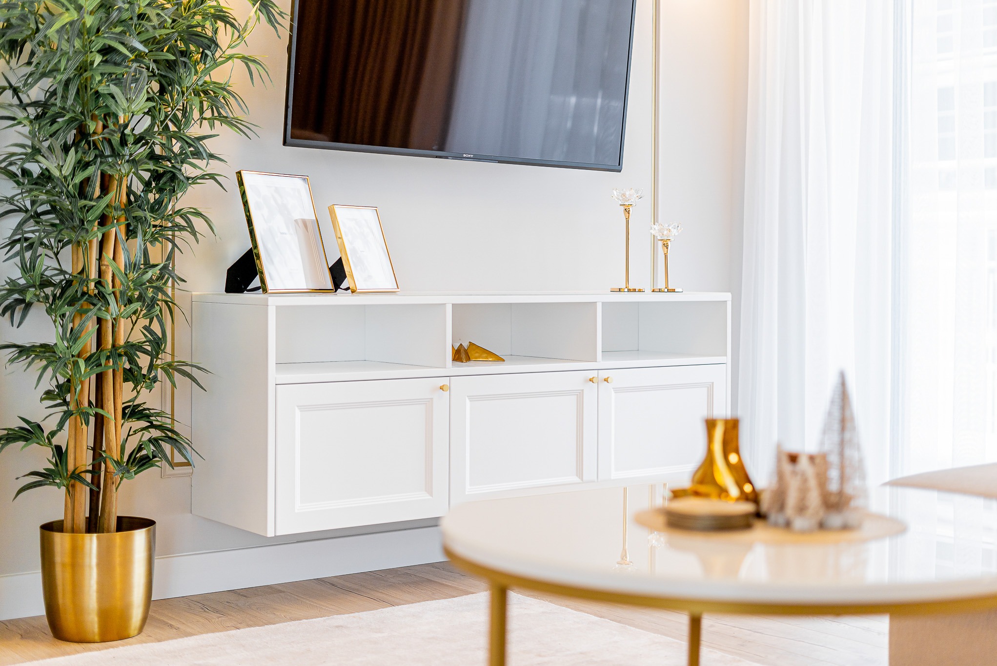 Jasne wnętrze: biała szafka RTV z dekoracjami (ramki, świeczniki), telewizor na ścianie, roślina w złotej donicy i okrągły stolik. Minimalistyczny design.