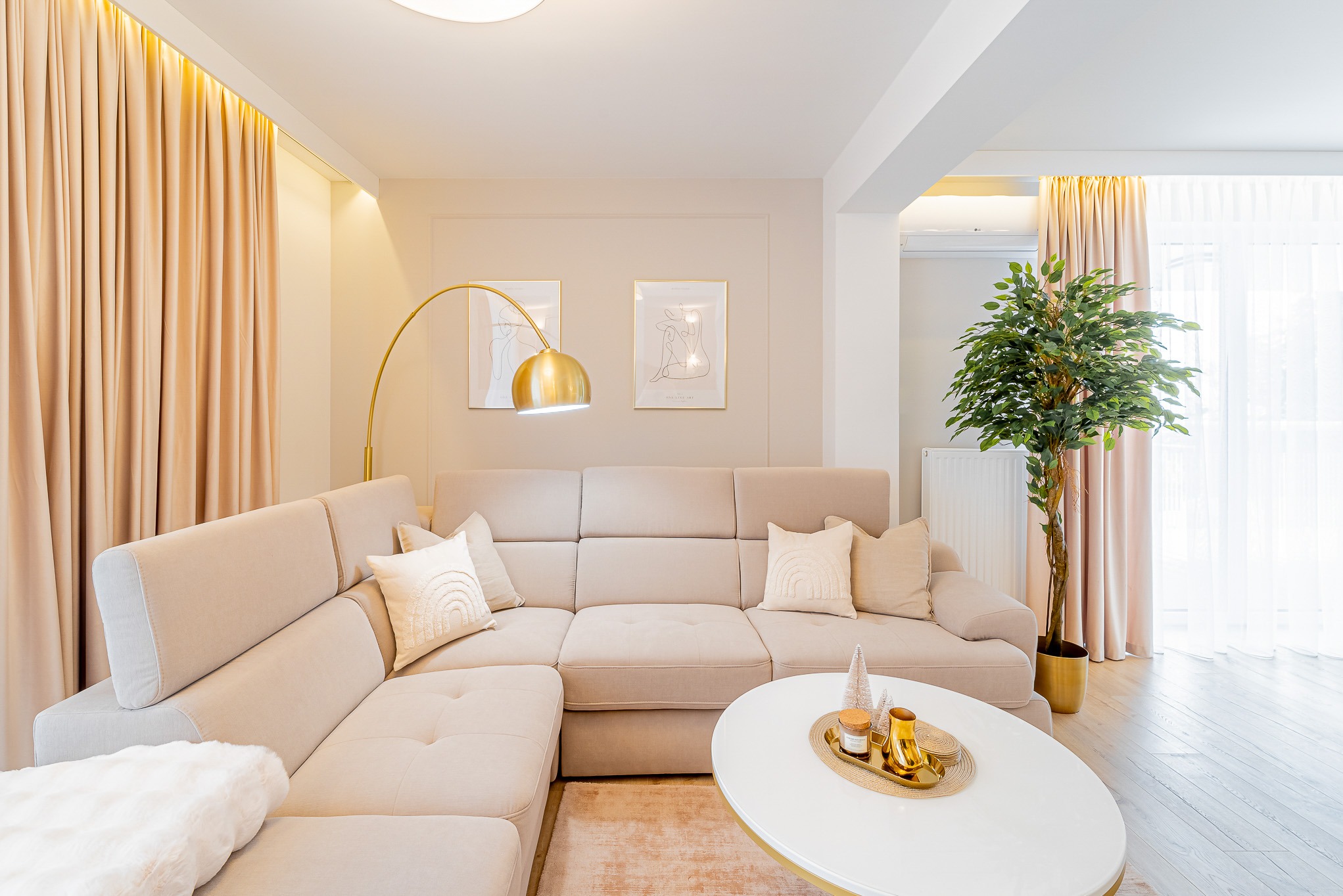 Jasne wnętrze salonu z narożną sofą, złotą lampą łukową i okrągłym stolikiem kawowym. Beżowe zasłony i minimalistyczne grafiki na ścianie dopełniają aranżację.