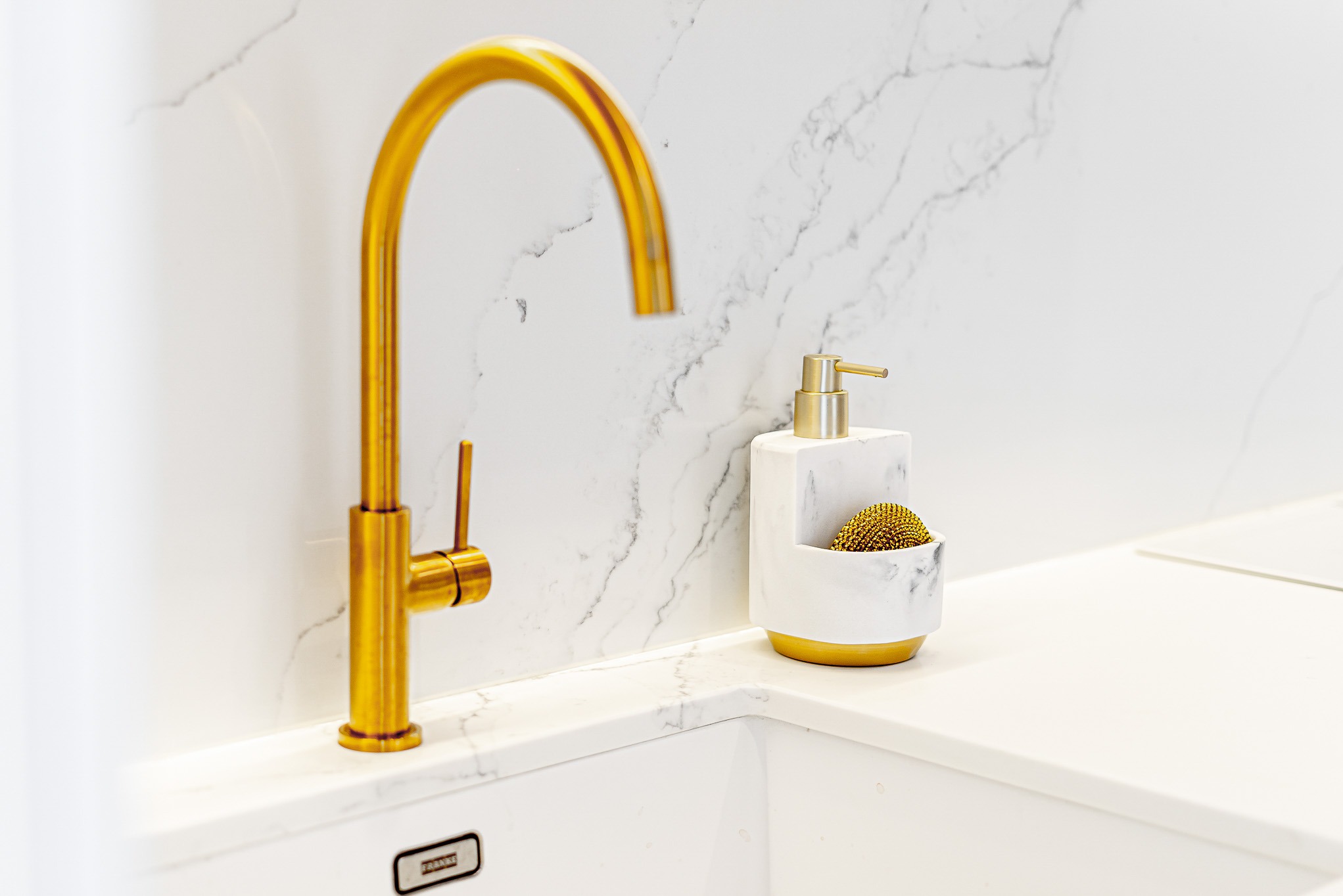 Błyszcząca, złota bateria kuchenna z dozownikiem na tle białego blatu z marmurowym wzorem. Minimalistyczny design, oświetlenie LED pod blatem.