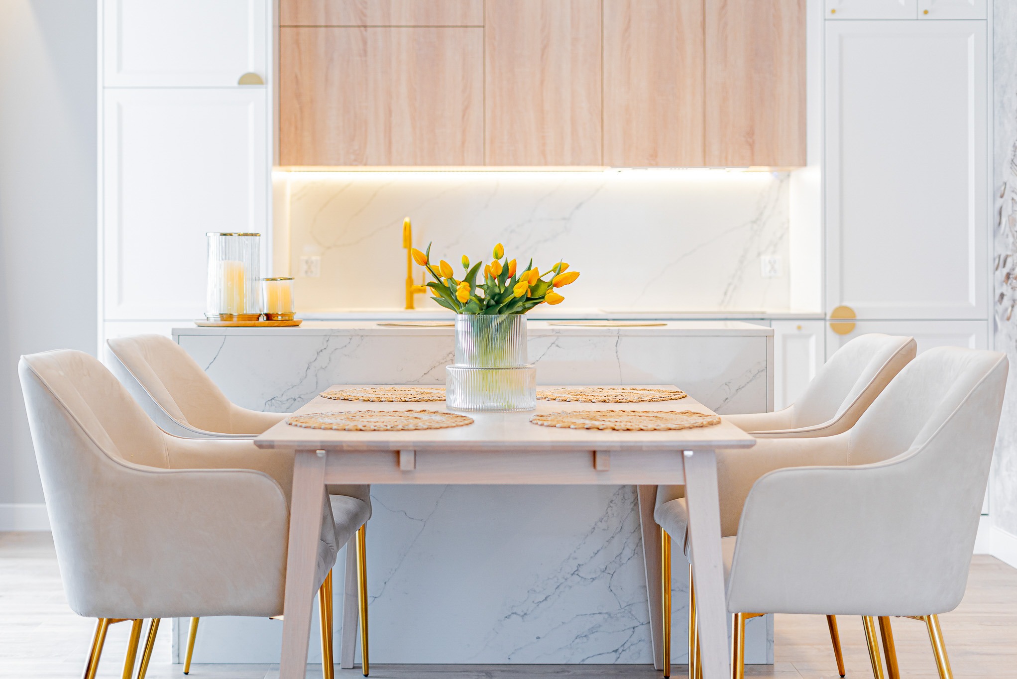 Jasna jadalnia z drewnianym stołem, beżowymi krzesłami na złotych nogach i bukietem żółtych tulipanów. W tle marmurowa wyspa kuchenna i białe szafki.