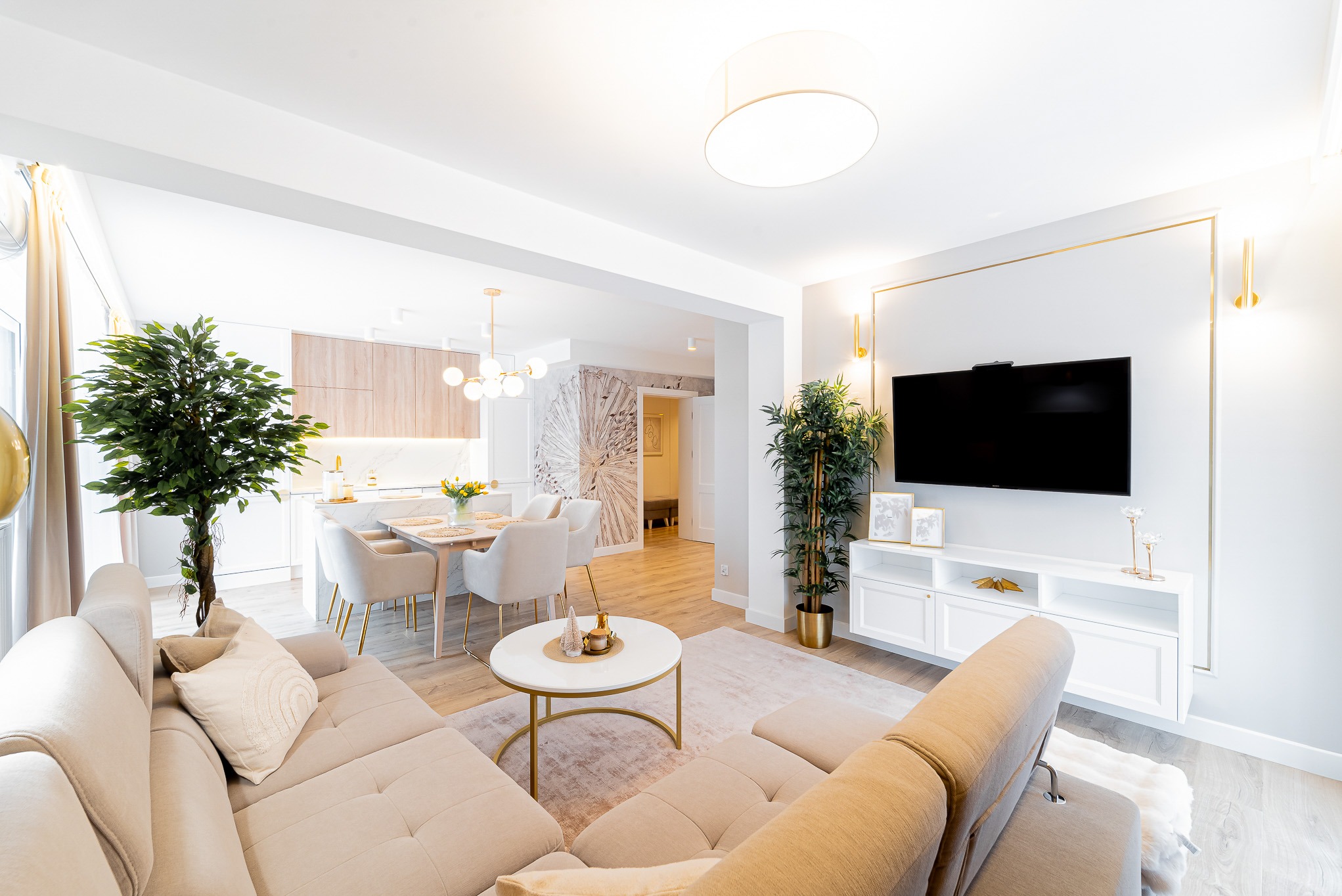 Jasne wnętrze salonu z aneksem kuchennym. Beżowa sofa narożna, biały stolik kawowy ze złotymi nogami, telewizor na ścianie z dekoracyjną ramą. Nowoczesny design.