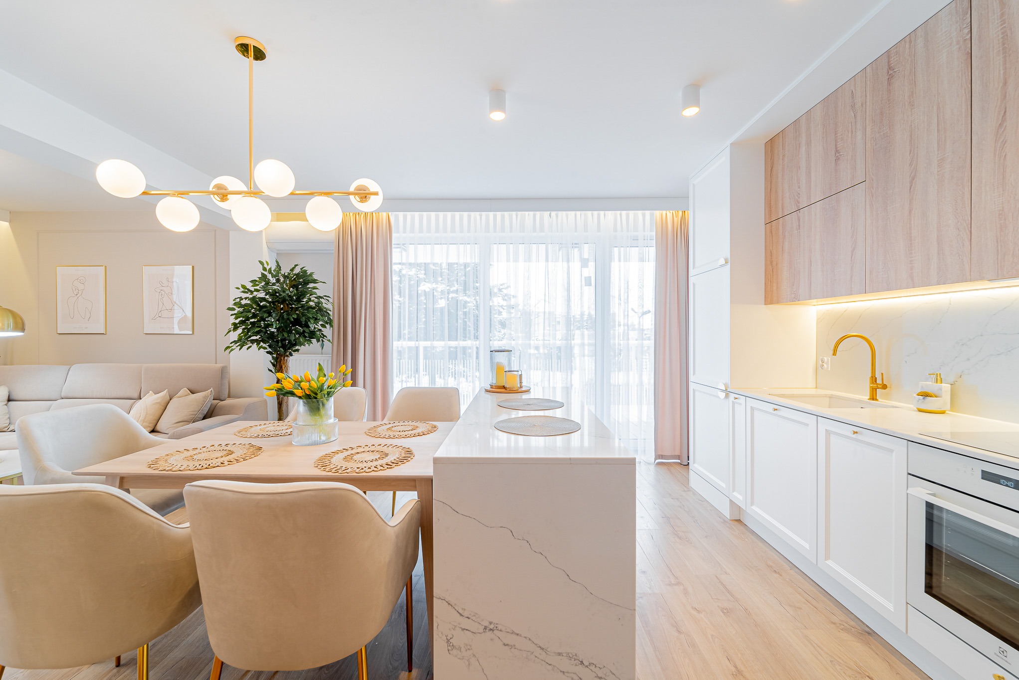 Jasne wnętrze: stół z żółtymi tulipanami, krzesła z weluru, marmurowy blat kuchenny, złota armatura i lampa. W tle sofa i grafiki na ścianie.