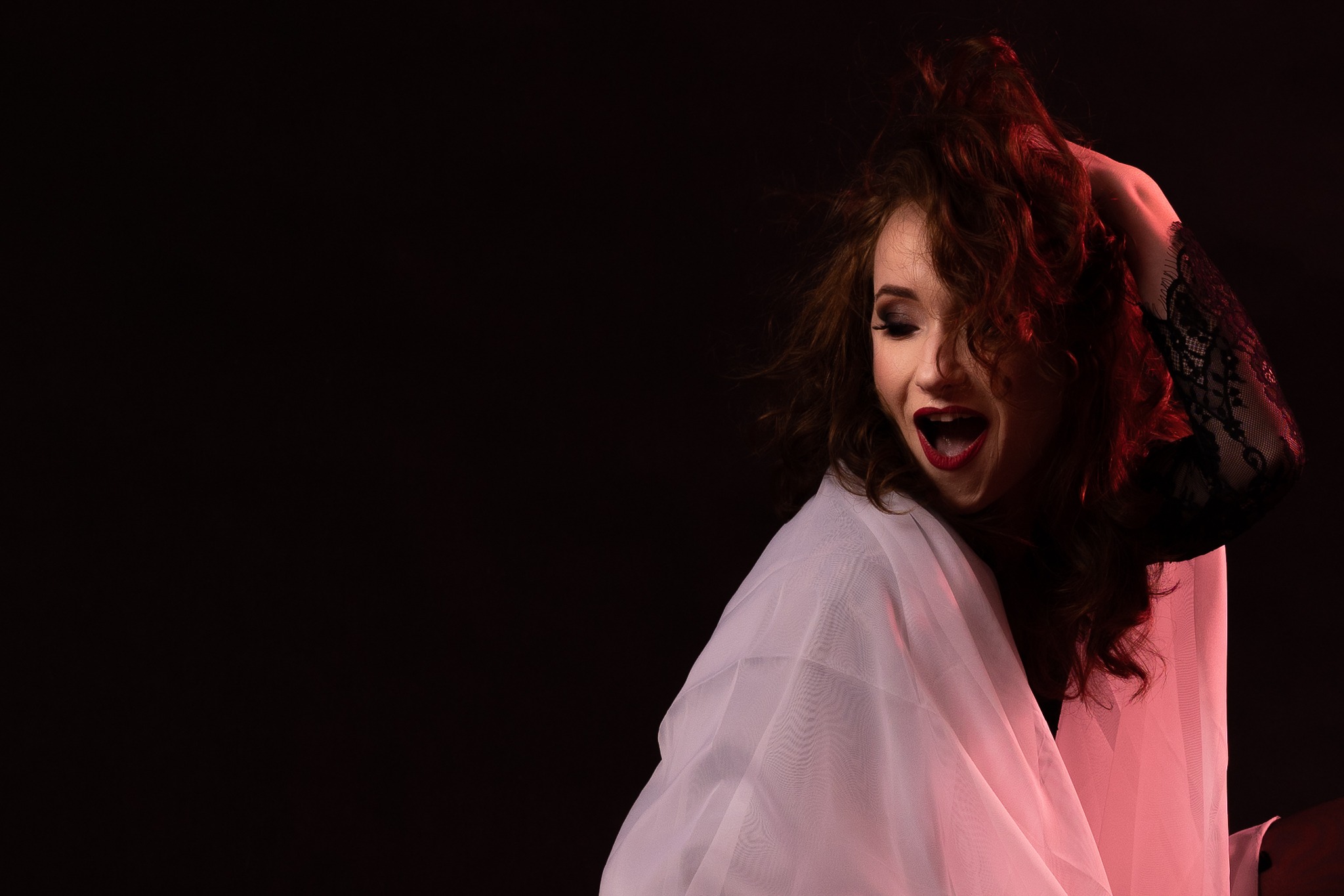 Portret kobiety w białej, zwiewnej tkaninie i czarnej koronkowej rękawiczce, z rozpuszczonymi, rudymi włosami, odchyloną głową i otwartymi ustami na ciemnym tle.