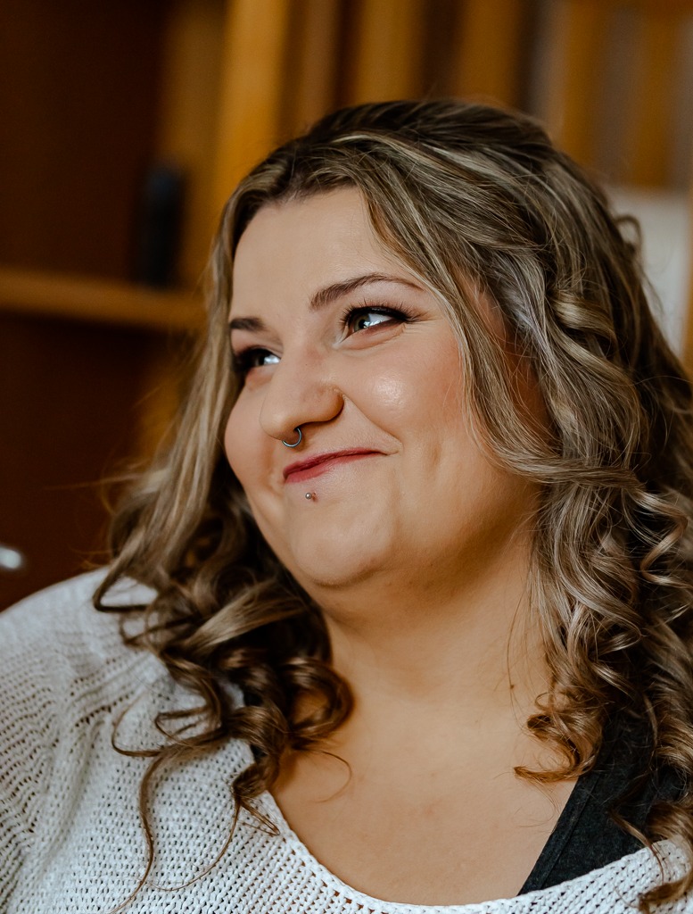 Portret uśmiechniętej kobiety z piercingiem w nosie i na brodzie, z blond włosami w lokach, ubrana w biały sweter o grubym splocie na tle drewnianych półek.