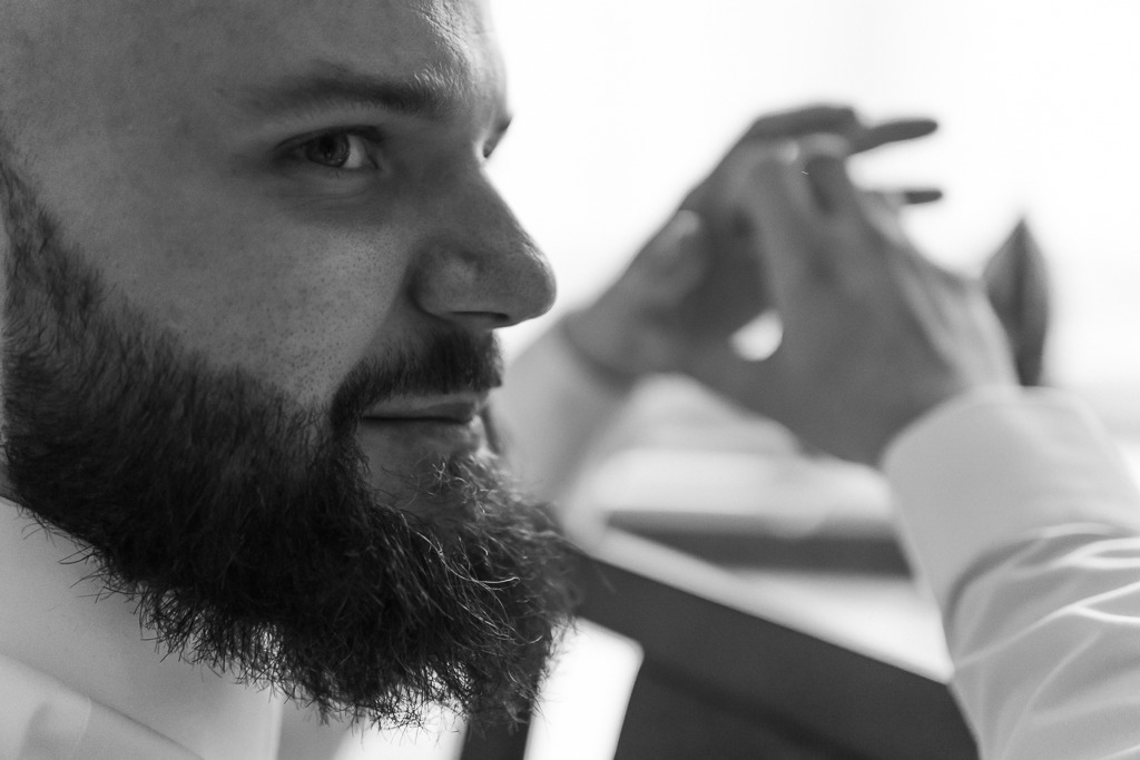 Portret mężczyzny z brodą w białej koszuli, skierowany w lewo, z dłońmi w tle w nieostrym ujęciu.