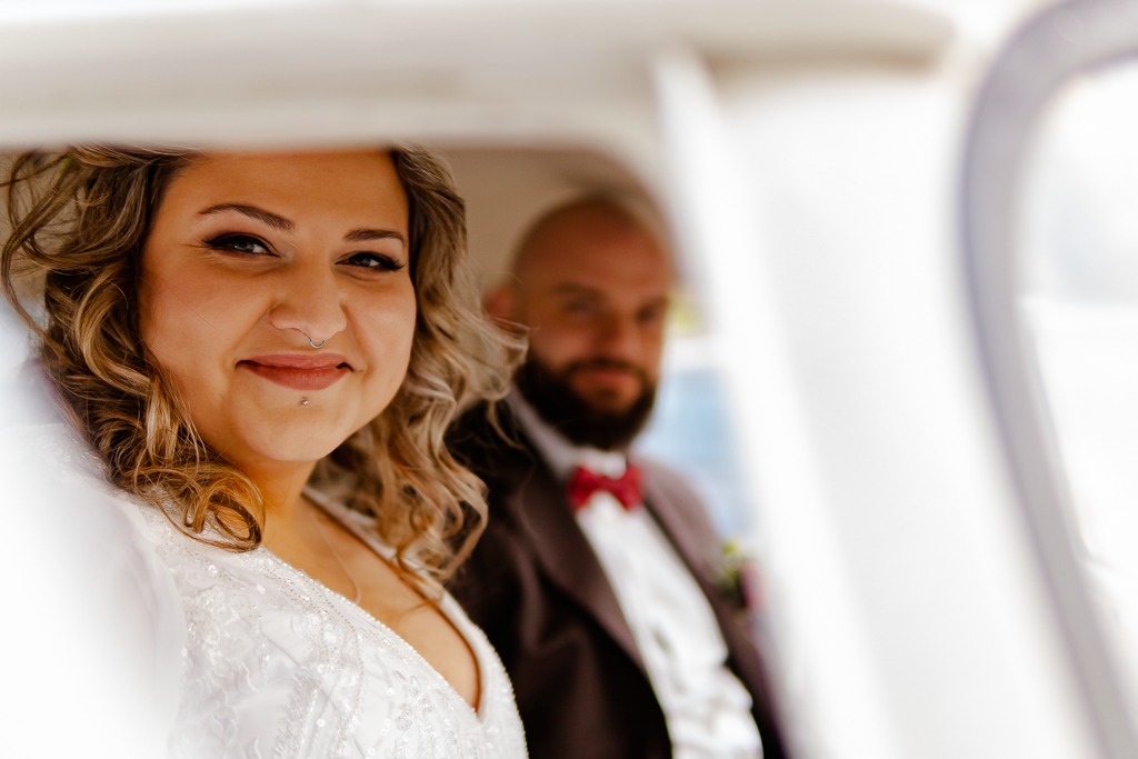 Uśmiechnięta panna młoda z kolczykiem w przegrodzie nosowej i na brodzie, w białej sukni z koronką, obok niewyraźny pan młody z czerwoną muszką w tle.