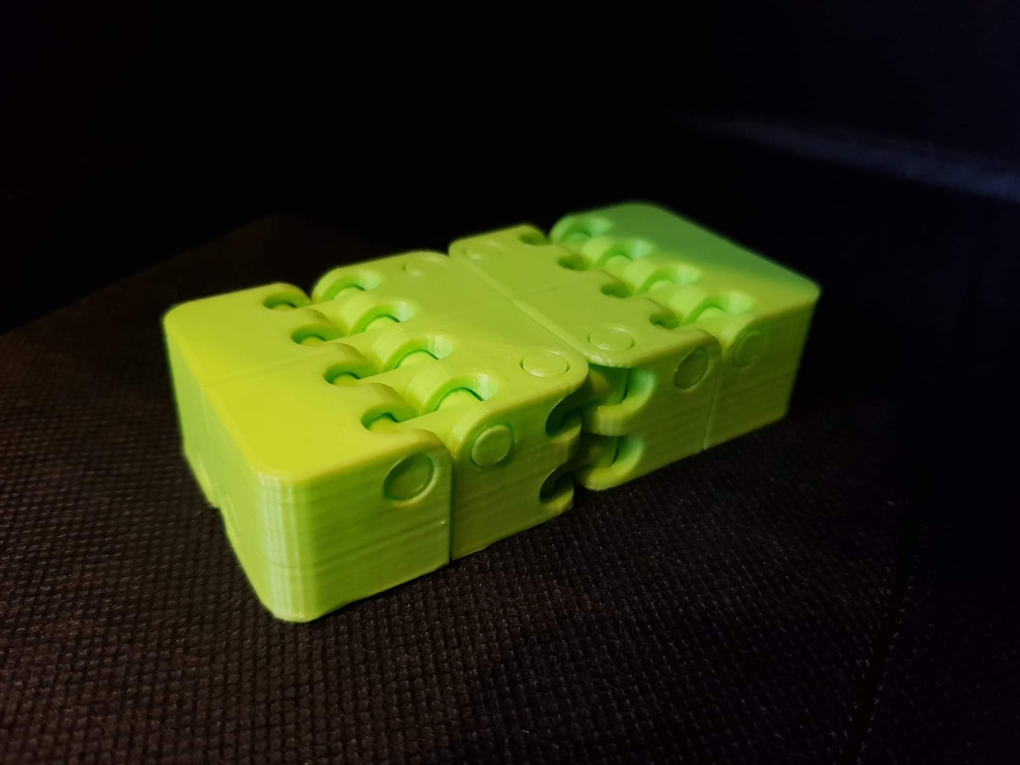 Jasnozielony, przegubowy model 3D z otworami, wydrukowany z widocznymi warstwami, leżący na ciemnej, teksturowanej powierzchni.