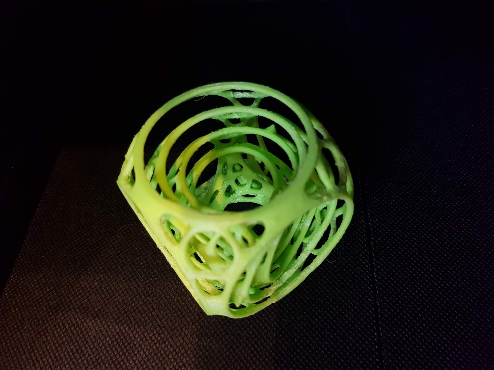 Zielony, ażurowy model 3D w kształcie sześcianu z okrągłymi otworami, wydrukowany na drukarce 3D, leżący na czarnej, teksturowanej powierzchni.