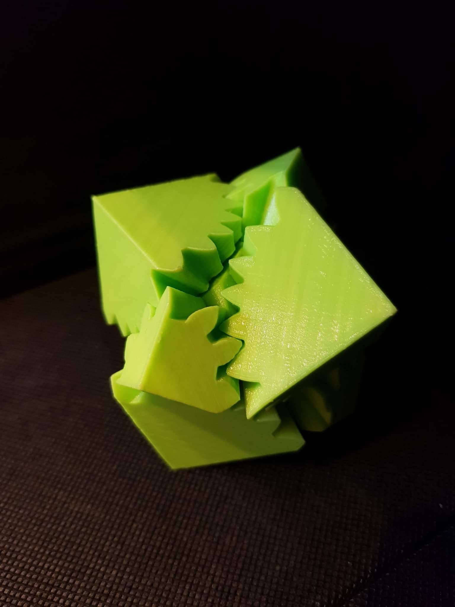 Jasnozielony, trójwymiarowy model kostki z wypustkami, wydrukowany na drukarce 3D, leżący na ciemnej powierzchni. Widoczne ślady warstw wydruku.