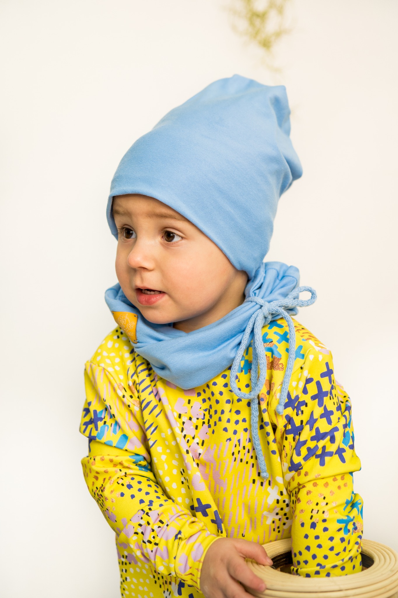 Mały chłopiec w błękitnej czapce i kominie, ubrany w żółtą bluzę z kolorowym wzorem, trzymający drewnianą zabawkę na tle białej ściany, patrzy w bok.