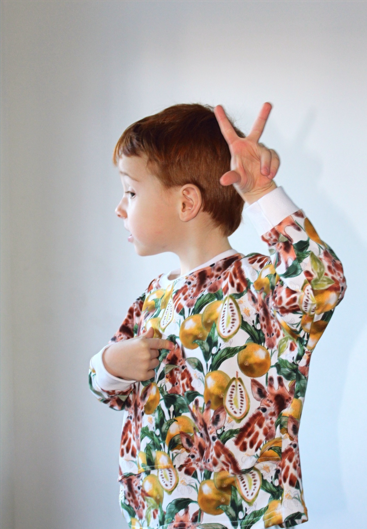 Chłopiec z rudymi włosami w bluzie z nadrukiem żyraf, cytryn i przekrojonych gruszek, pokazujący palcem na bluzę i gest 'V' dłonią.