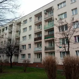 Wielorodzinny budynek po ociepleniu elewacji, z balkonami i widocznymi klimatyzatorami, otoczony zielenią i drzewami bez liści.