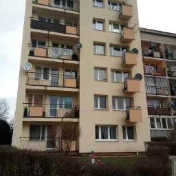 Wielopiętrowy budynek mieszkalny z balkonami i antenami satelitarnymi, widok z poziomu gruntu, krzewy na pierwszym planie.