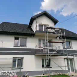 Dwupiętrowy dom z balkonami po tynkowaniu elewacji, z widocznymi rusztowaniami i anteną satelitarną na szczycie dachu, sfotografowany w słoneczny dzień.