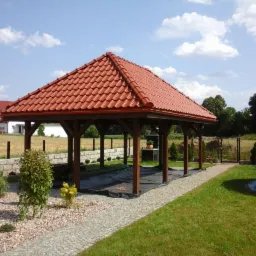 Zadaszona altana ogrodowa z czerwoną dachówką ceramiczną, widoczna z perspektywy ogrodu, z fragmentem ścieżki z kostki brukowej i basenem w tle.