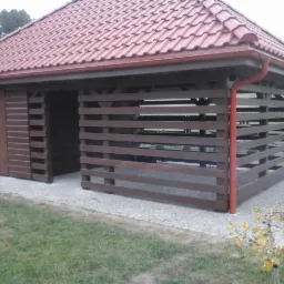 Zadaszona drewniana altana z czerwonymi rynnami i brązowymi elementami konstrukcyjnymi, położona na trawniku z kamienną ścieżką.