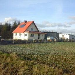 Żółty dom jednorodzinny z czerwoną dachówką i panelami słonecznymi na dachu, widziany z perspektywy pola w pochmurny dzień.