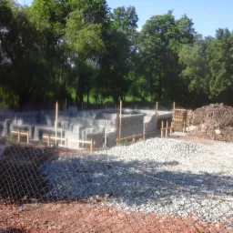 Wykop pod fundamenty domu w trakcie wznoszenia, widoczne bloczki betonowe, drewniane szalunki, hałda ziemi i gruzu, siatka ogrodzeniowa na pierwszym planie.