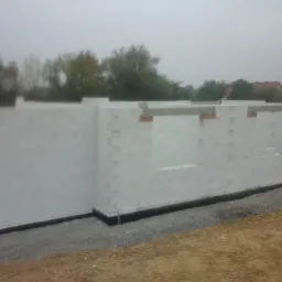 Budowa domu z białych bloczków: wzniesione ściany fundamentowe z widoczną izolacją przeciwwilgociową, częściowo wypełnione zaprawą, na tle pochmurnego nieba i zieleni.