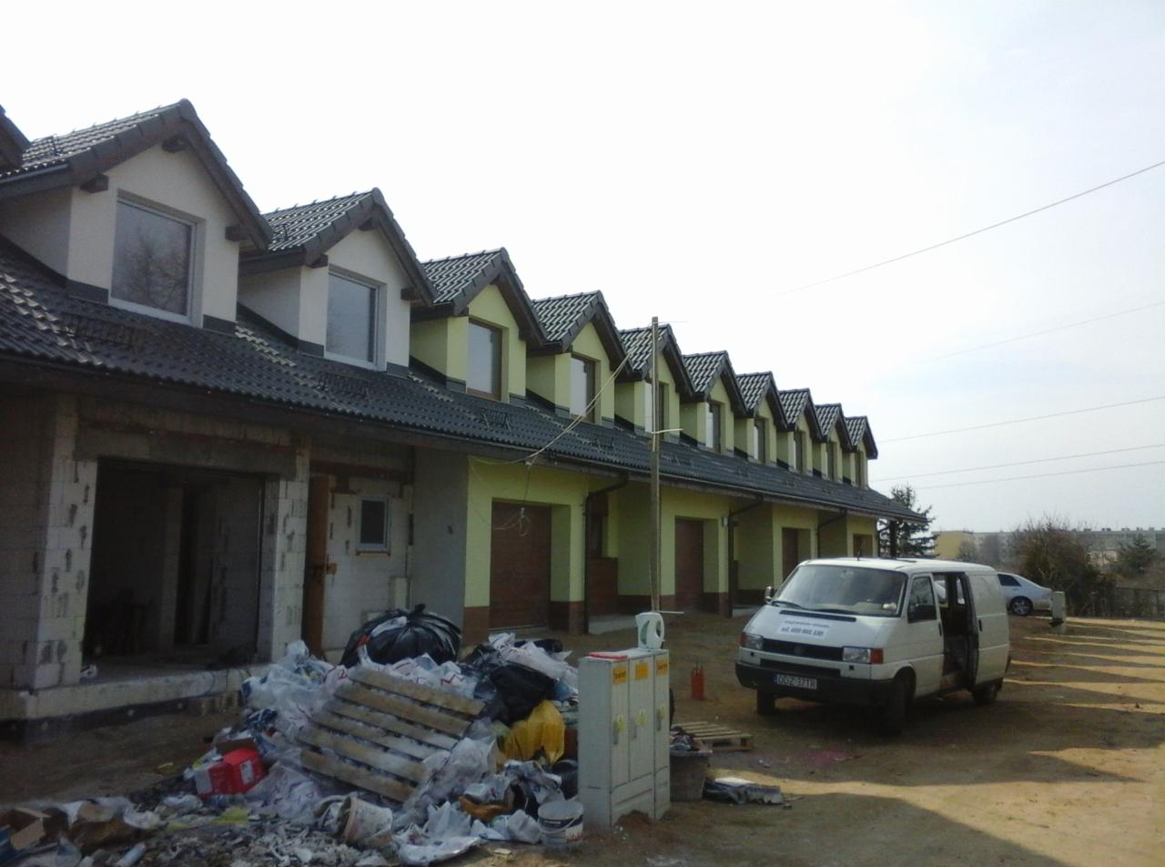 Szeregówka w trakcie budowy, widoczne niezamknięte garaże, śmieci budowlane przed budynkiem, zaparkowany biały bus.