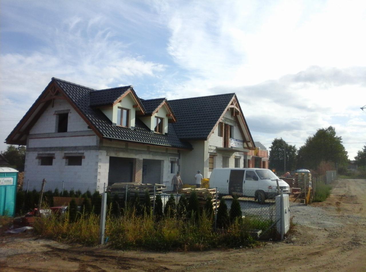 Surowy stan domu murowanego z ciemnym dachem, widoczna furgonetka, palety z materiałami budowlanymi i przenośna toaleta na placu budowy.