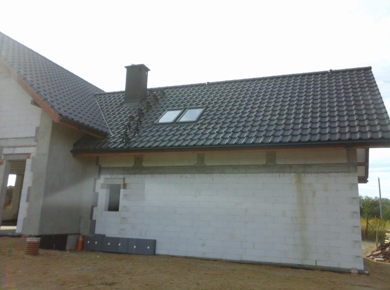 Dom w trakcie budowy z częściowo ukończonym dachem z dachówki ceramicznej, widoczne okna dachowe i komin, ściany z białego bloczka, przygotowane elementy do dalszej budowy.