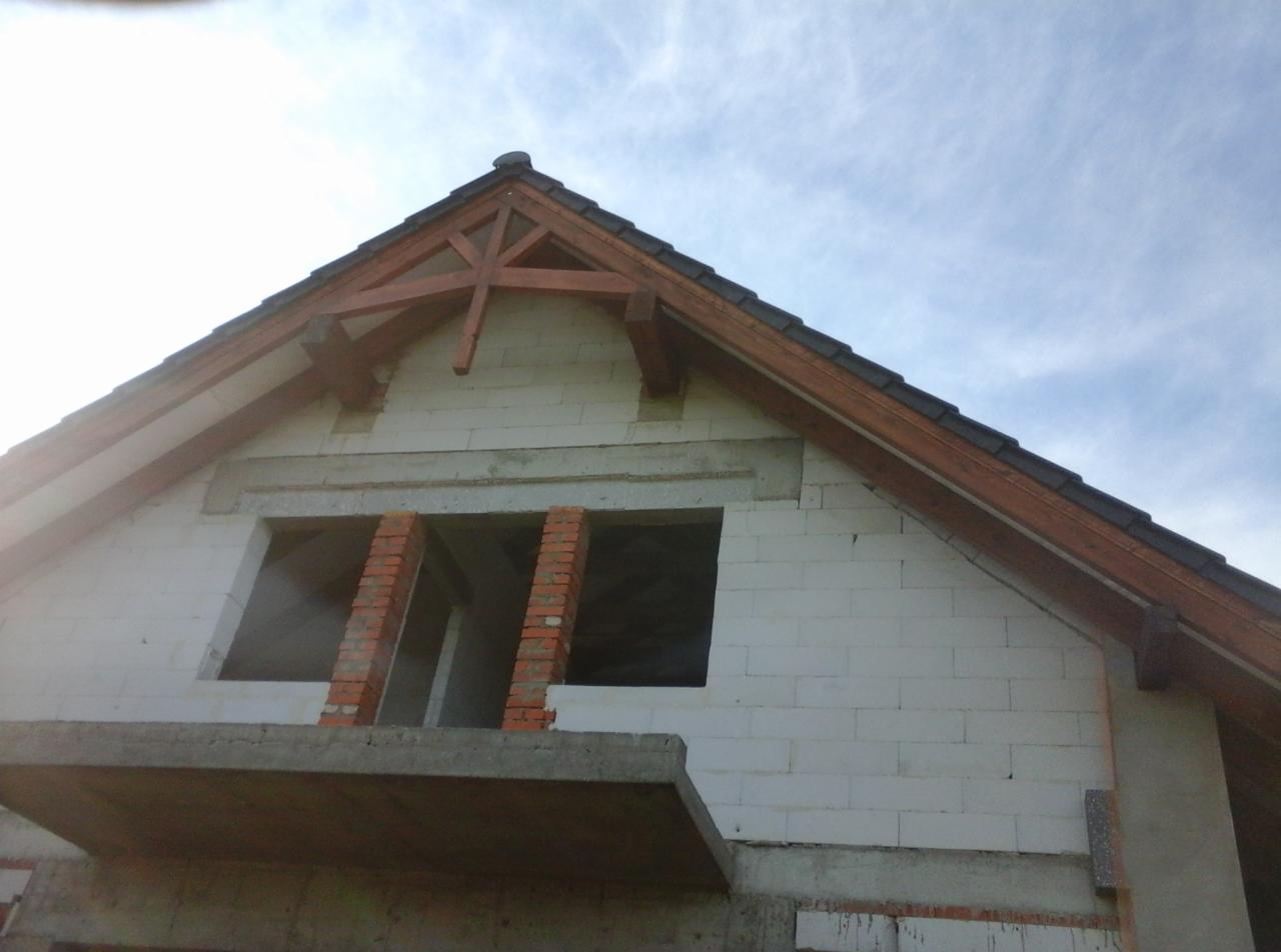 Surowy stan domu z białych bloczków, widoczna więźba dachowa z ciemnego drewna, niezadaszony balkon i fragmenty murowanej cegły wokół okien, na tle błękitnego nieba.