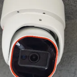 Nowoczesna, biała kamera obrotowa z pomarańczowym akcentem, zamontowana pod szarą, chropowatą powierzchnią sufitu.