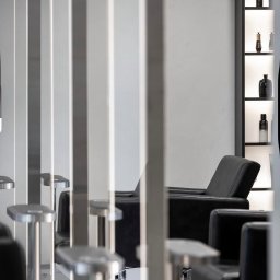 F I X M A S T E R S - Nowoczesne fotele fryzjerskie w minimalistycznym salonie, widoczne przez metalowe przegrody. Na ścianie podświetlone półki z kosmetykami.
