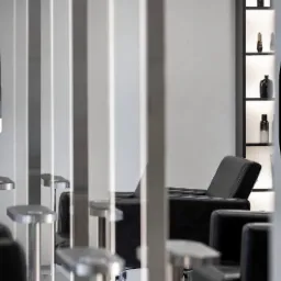 Nowoczesne fotele fryzjerskie w minimalistycznym salonie, widoczne przez metalowe przegrody. Na ścianie podświetlone półki z kosmetykami.