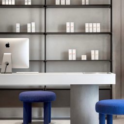 F I X M A S T E R S - Minimalistyczne wnętrze: białe biurko z komputerem Apple, kobaltowe pufy i półki z produktami kosmetycznymi na szarym tle. Nowoczesny design.