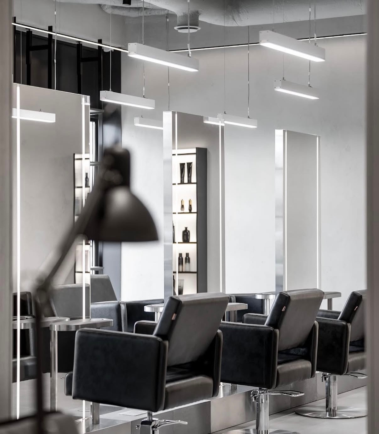 Nowoczesne wnętrze salonu fryzjerskiego z czarnymi fotelami, lustrami z oświetleniem LED i minimalistycznym designem. Widoczne półki z kosmetykami i lampa biurkowa.