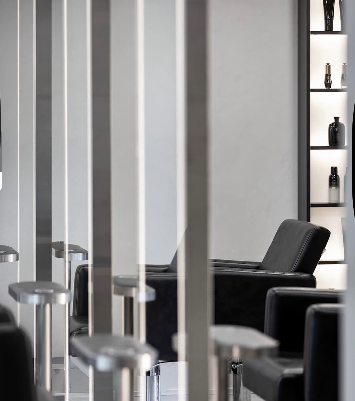 Nowoczesne fotele fryzjerskie w minimalistycznym salonie, widoczne przez metalowe przegrody. Na ścianie podświetlone półki z kosmetykami.