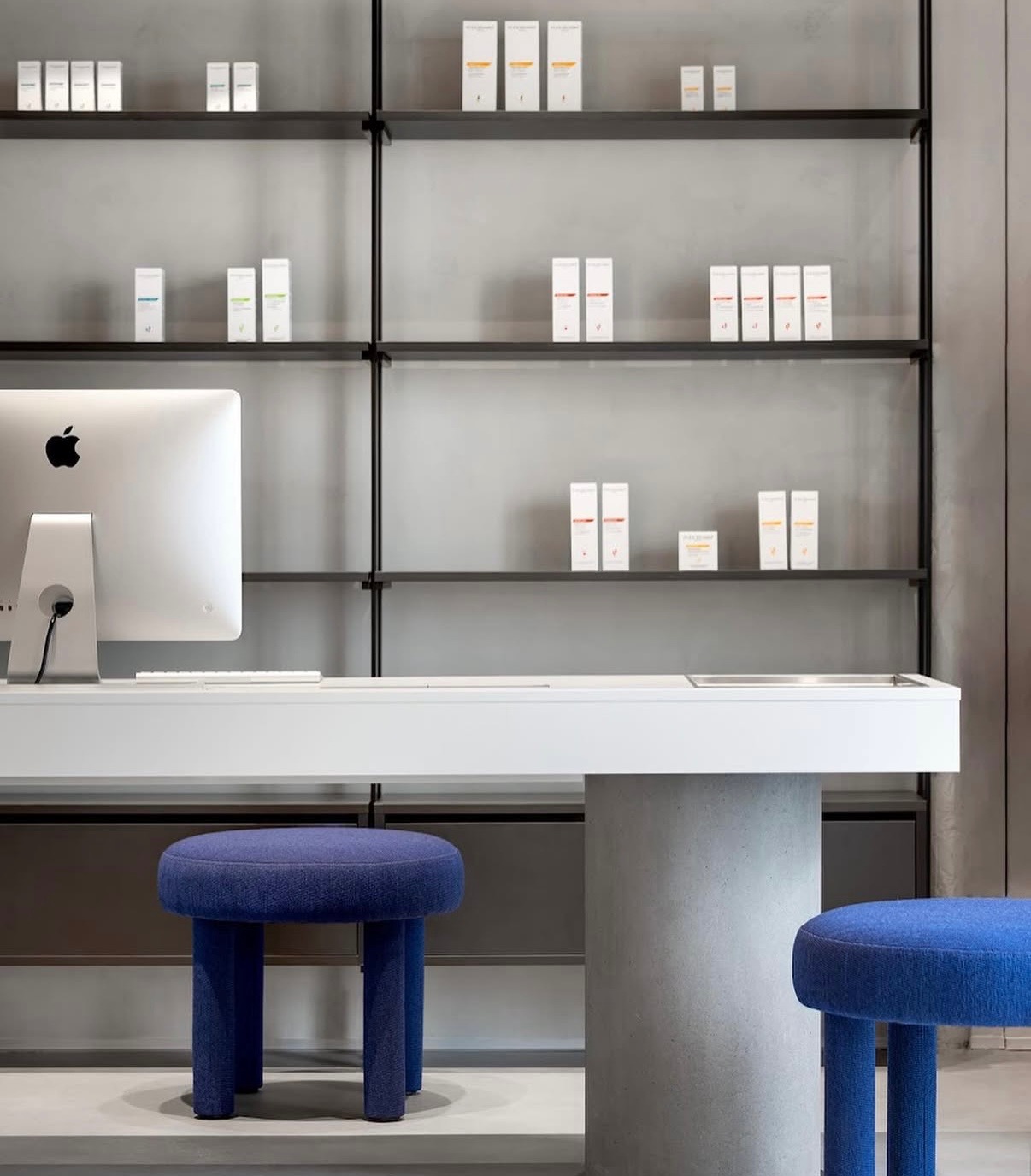 Minimalistyczne wnętrze: białe biurko z komputerem Apple, kobaltowe pufy i półki z produktami kosmetycznymi na szarym tle. Nowoczesny design.