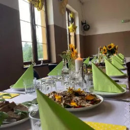 Uroczysty stół nakryty białym obrusem, dekorowany zielonymi serwetkami, kwiatami słonecznika i świecą, z balonami w tle, przygotowany na przyjęcie.